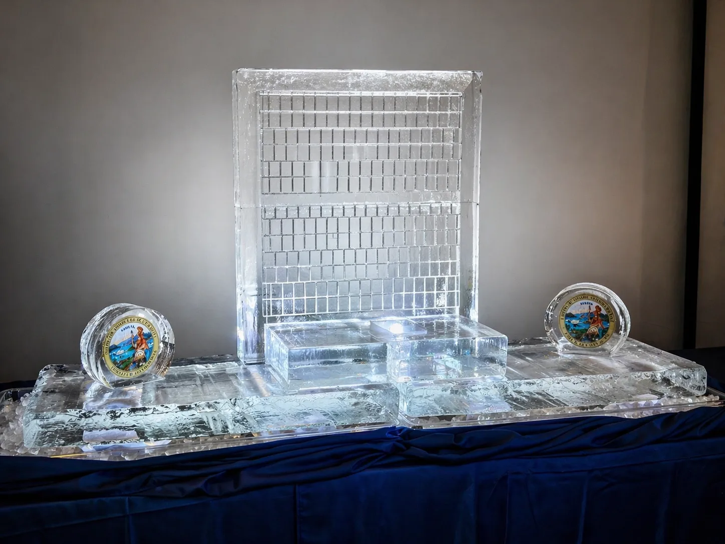 Large rectangular ice sculpture depicting a gridded building facade flanked by two circular ice-embedded official seals on a blue draped table.