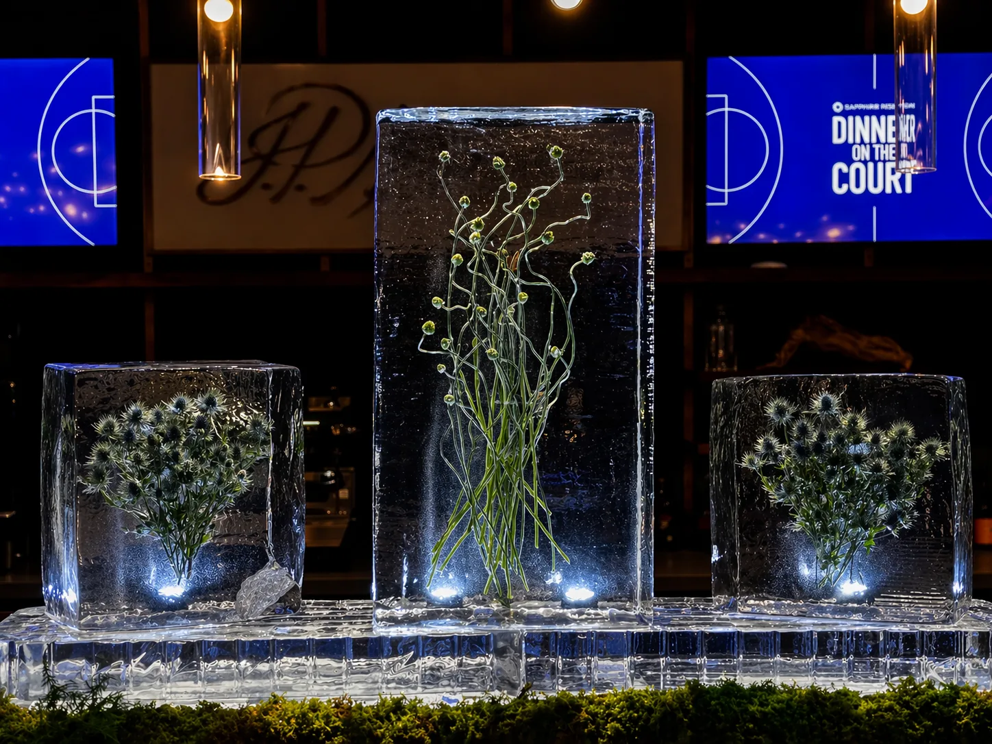 Three clear ice blocks with embedded flowers and branches on a tiered ice platform, blue-lit, with Dinner on the Court signage visible behind.