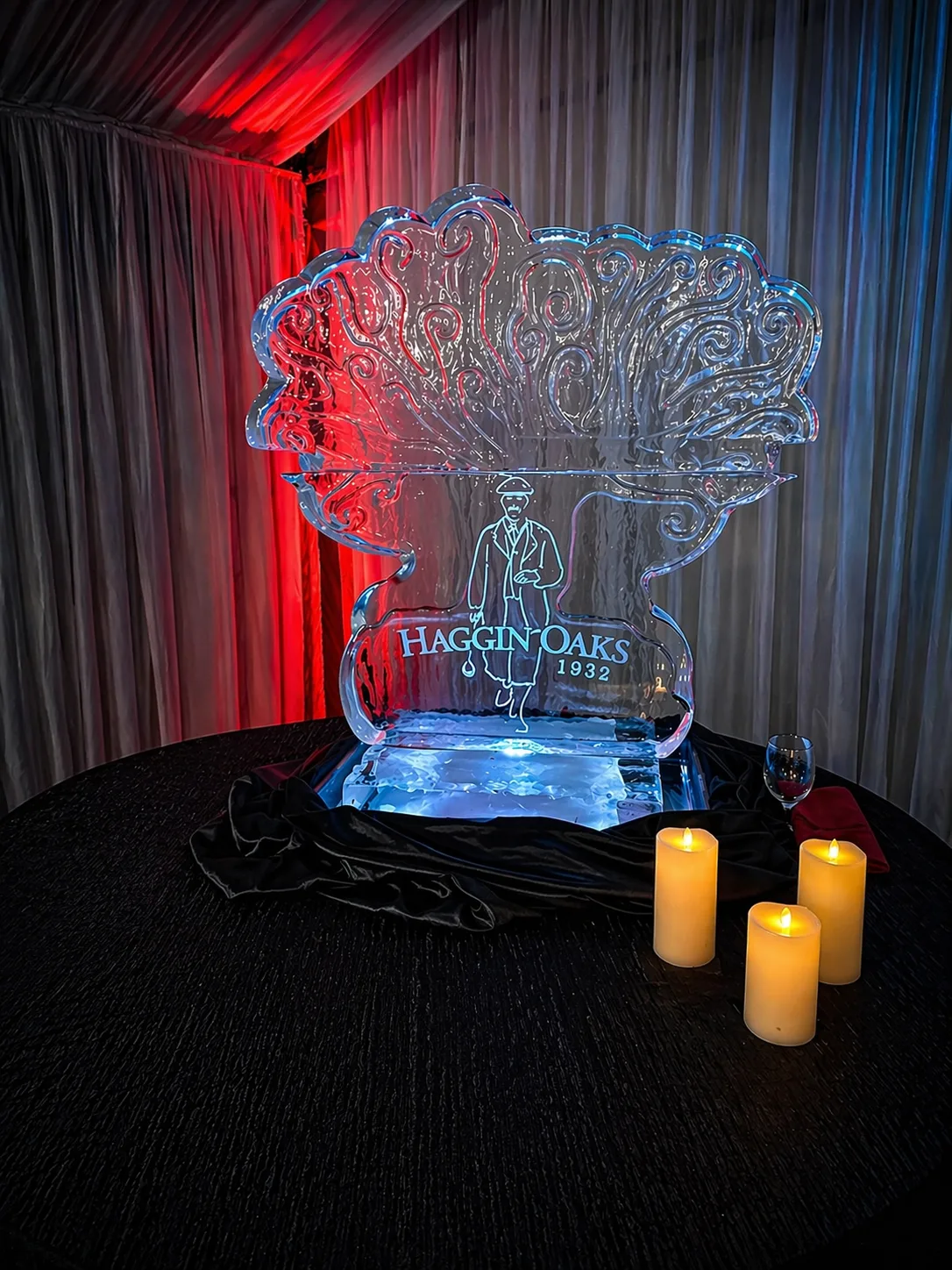 Ice sculpture engraved with HAGGIN OAKS 1932 and a golfer figure beneath a decorative tree, blue-lit on a black draped table with candles.
