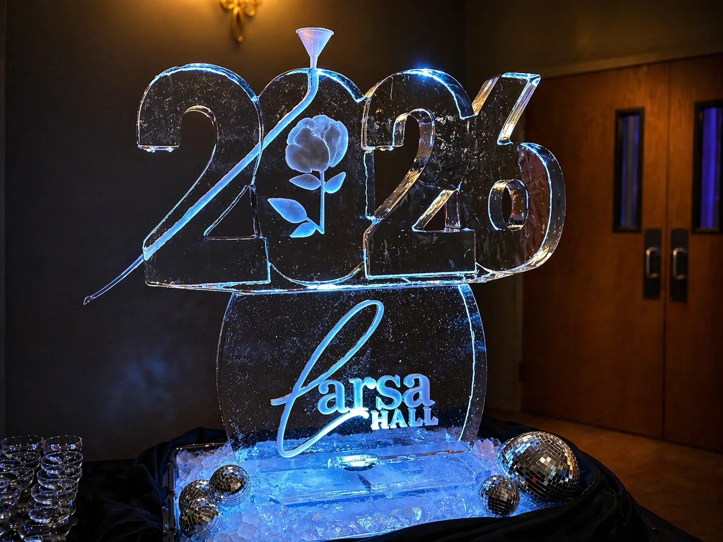 Large clear ice sculpture carved with 2026 numerals and a rose, displaying the text Larsa HALL in script, blue-lit on a black draped table.