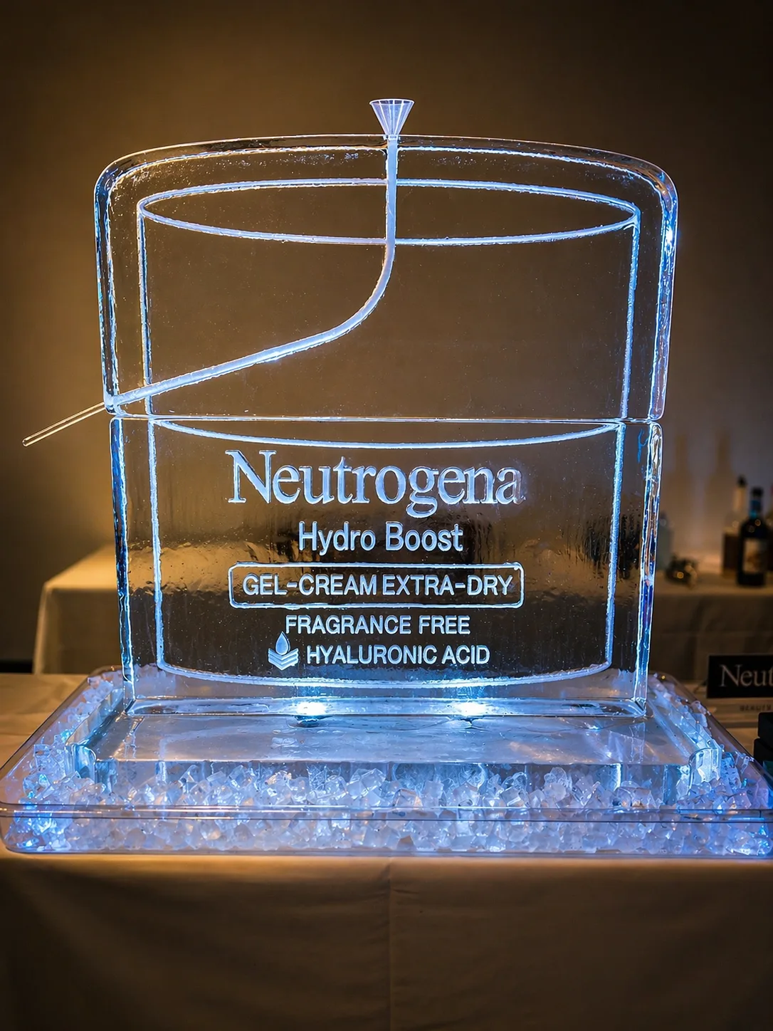 Clear ice sculpture carved to resemble a Neutrogena Hydro Boost Gel-Cream Extra-Dry jar with engraved product text, lit from below with blue light.