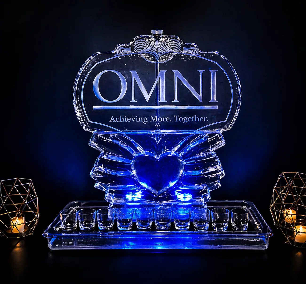 Ice sculpture carved with OMNI Achieving More. Together. logo, flanked by wing shapes and a centered heart, with a shot glass tray, lit blue.