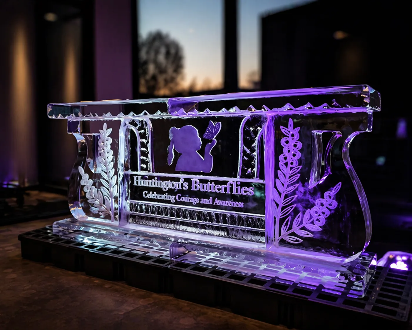 Ice bar structure engraved with Huntington's Butterflies Celebrating Courage and Awareness text, lit purple, at indoor evening event.
