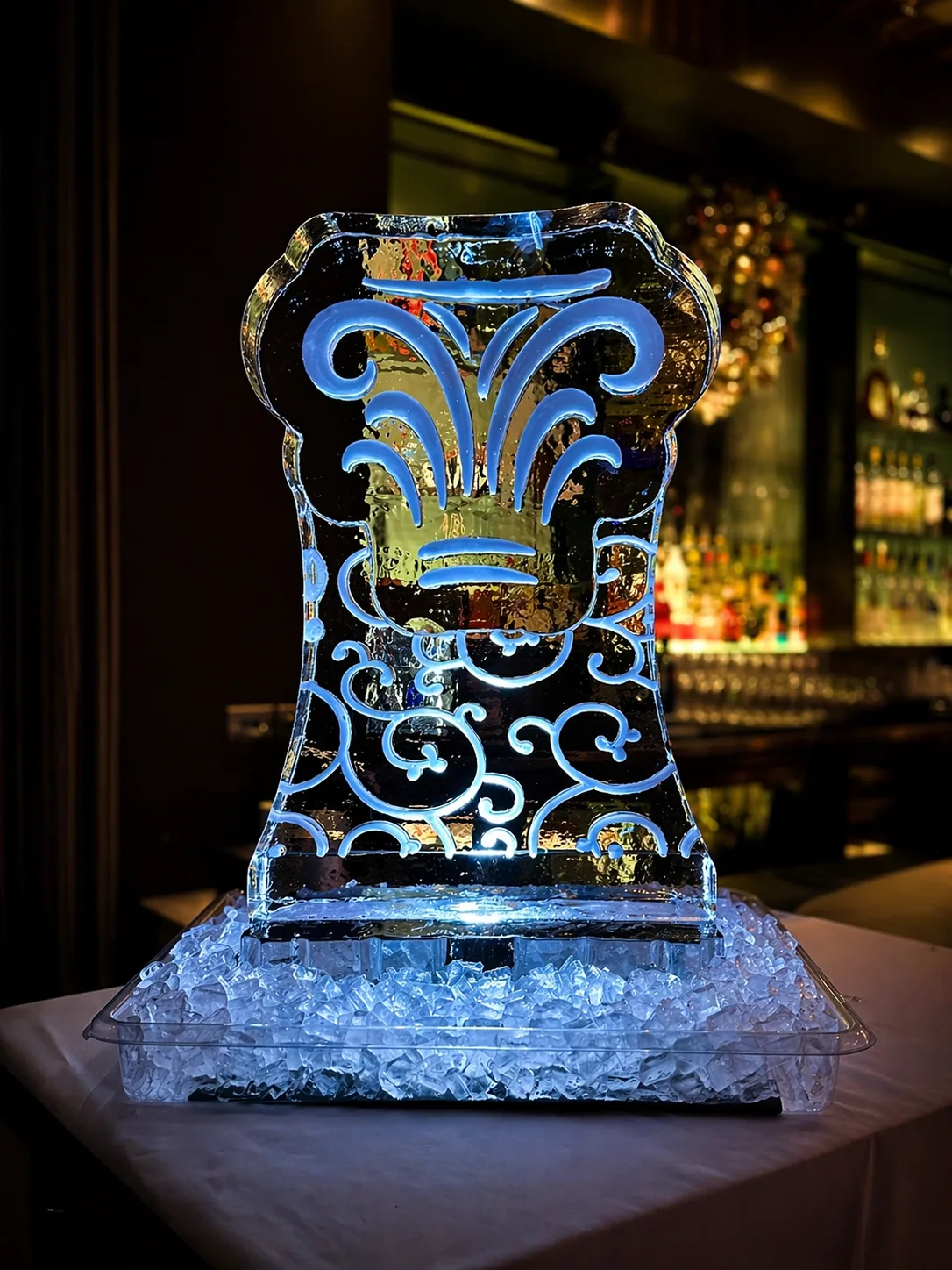 Large rectangular ice sculpture with carved blue-lit scrollwork and fleur-de-lis motif, displayed on crushed ice at a bar.