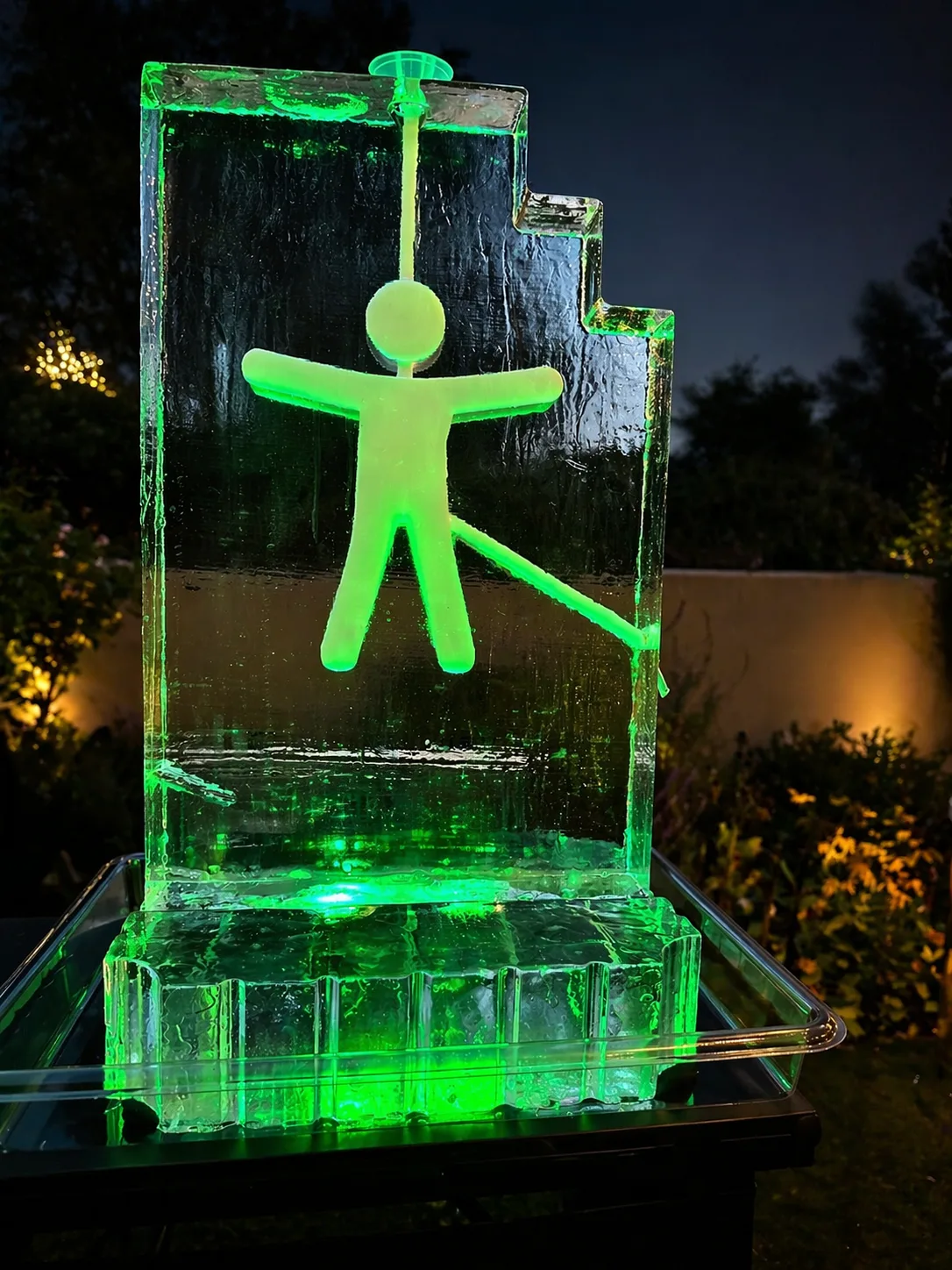 Large clear ice block with carved stick figure man glowing green, displayed outdoors at night on a raised base.