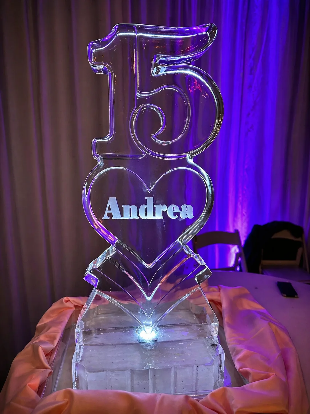 Tall clear ice sculpture displaying the number 15 above a heart engraved with the name Andrea, lit from below against a purple-lit draped backdrop.