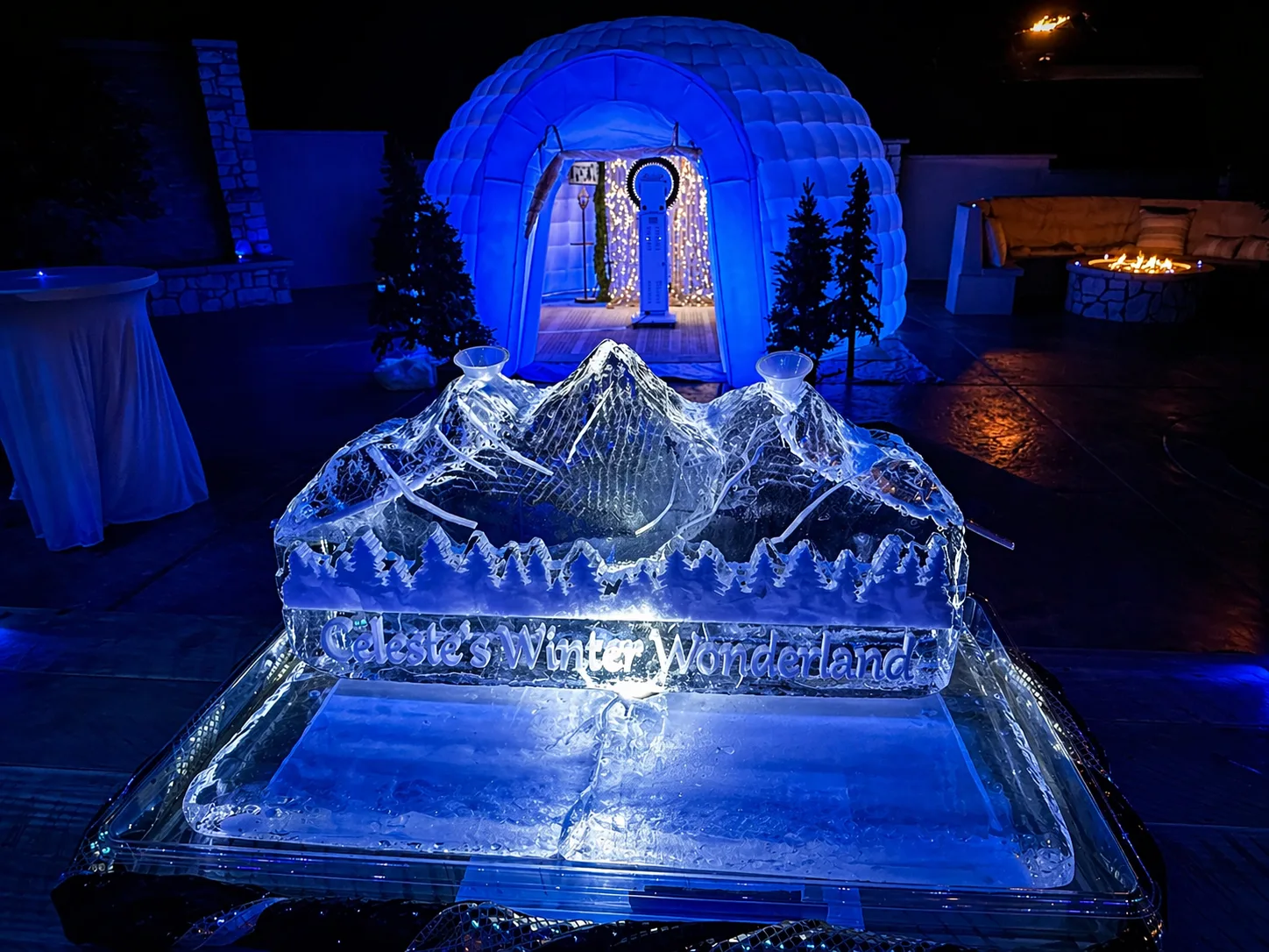 Large clear ice sculpture of mountain peaks with carved text reading Celeste's Winter Wonderland, set outdoors at night with blue-lit igloo in background.