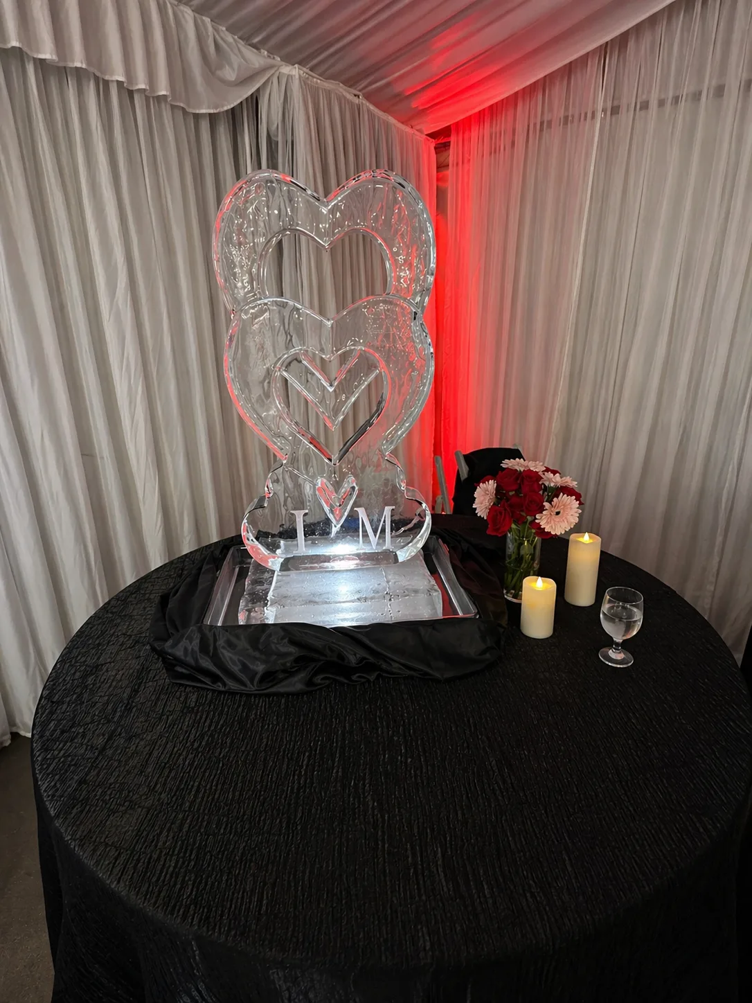 Tall clear ice sculpture of two nested hearts with monogram letters I and M, displayed on a black-draped table with red ambient lighting in background.