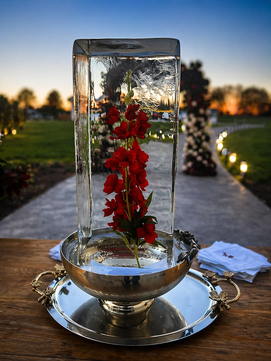 Clear rectangular ice block encasing a red gladiolus stem, displayed on a silver pedestal bowl at an outdoor evening wedding setting.
