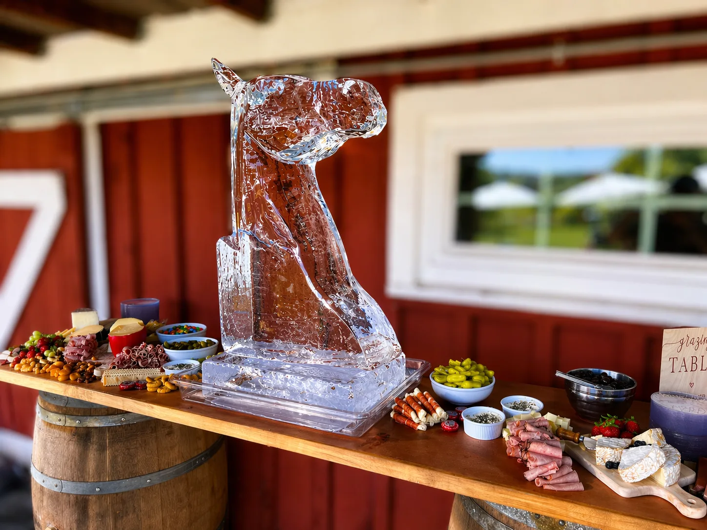 Clear ice horse head sculpture centered on a rustic wood plank table surrounded by charcuterie boards, olives, and fruit outside a red barn.