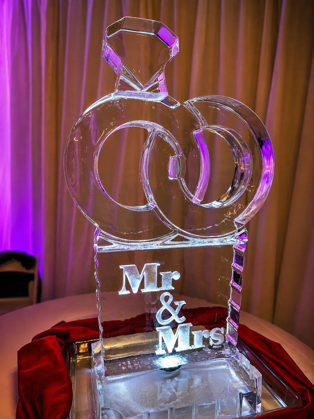 Clear ice sculpture of two interlocked wedding bands topped with a diamond ring carved with Mr & Mrs on the base, on a silver tray against a purple-lit backdrop.