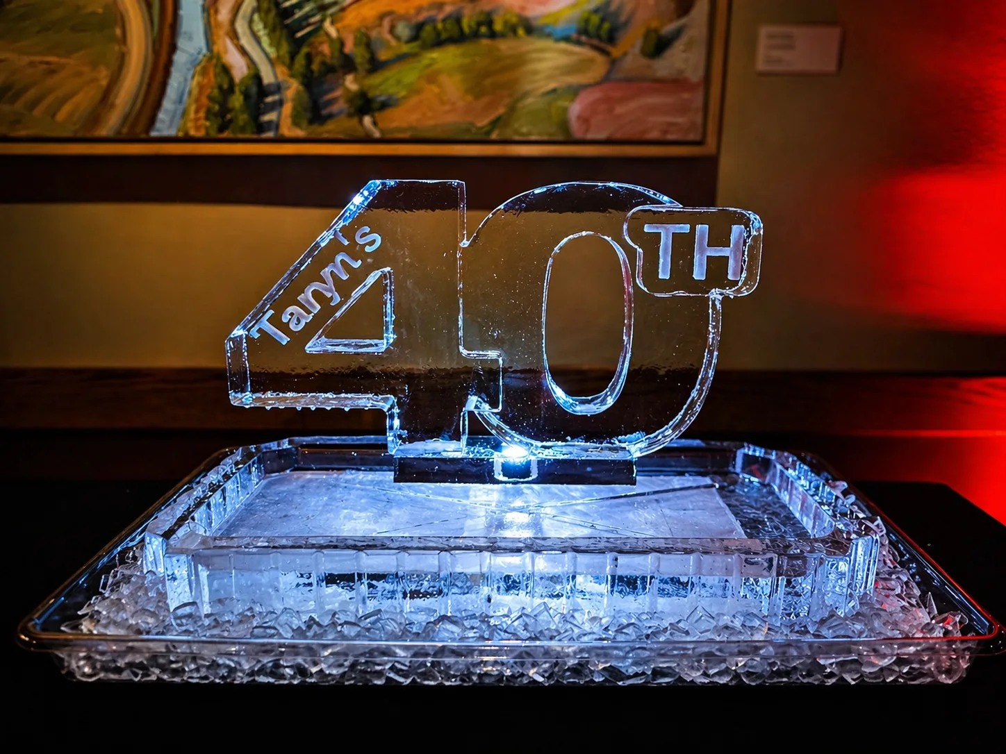 Clear ice sculpture carved with the text Taryn's 40TH displayed on a crushed ice tray, with blue underlighting and red ambient light in background.